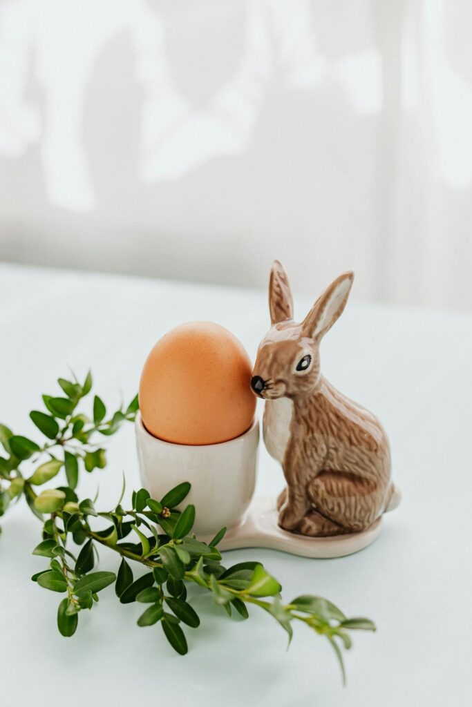 Jajko w beżowym kolorze spoczywa na białej porcelanowej podstawce, którą zdobi figura brązowego królika. Obok znajduje się gałązka z zielonymi liśćmi. Tło jest rozmyte w jasnych tonacjach.