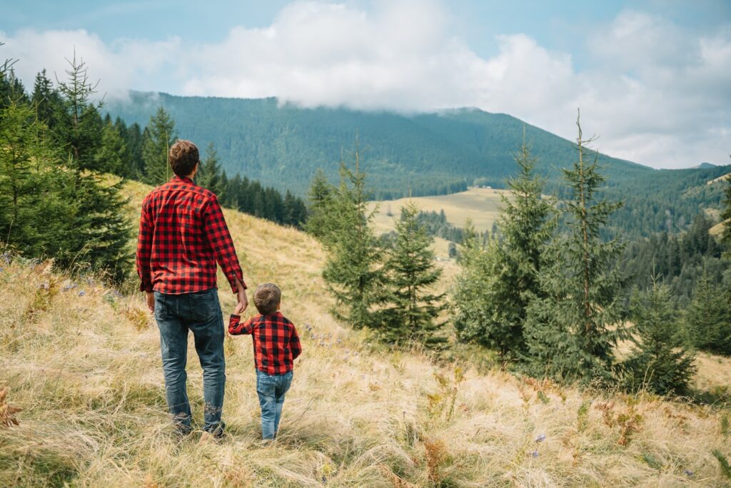 Szczęśliwy ojciec ze swoim młodym synem stoi tyłem na tle zielonego lasu, gór i nieba z chmurami.